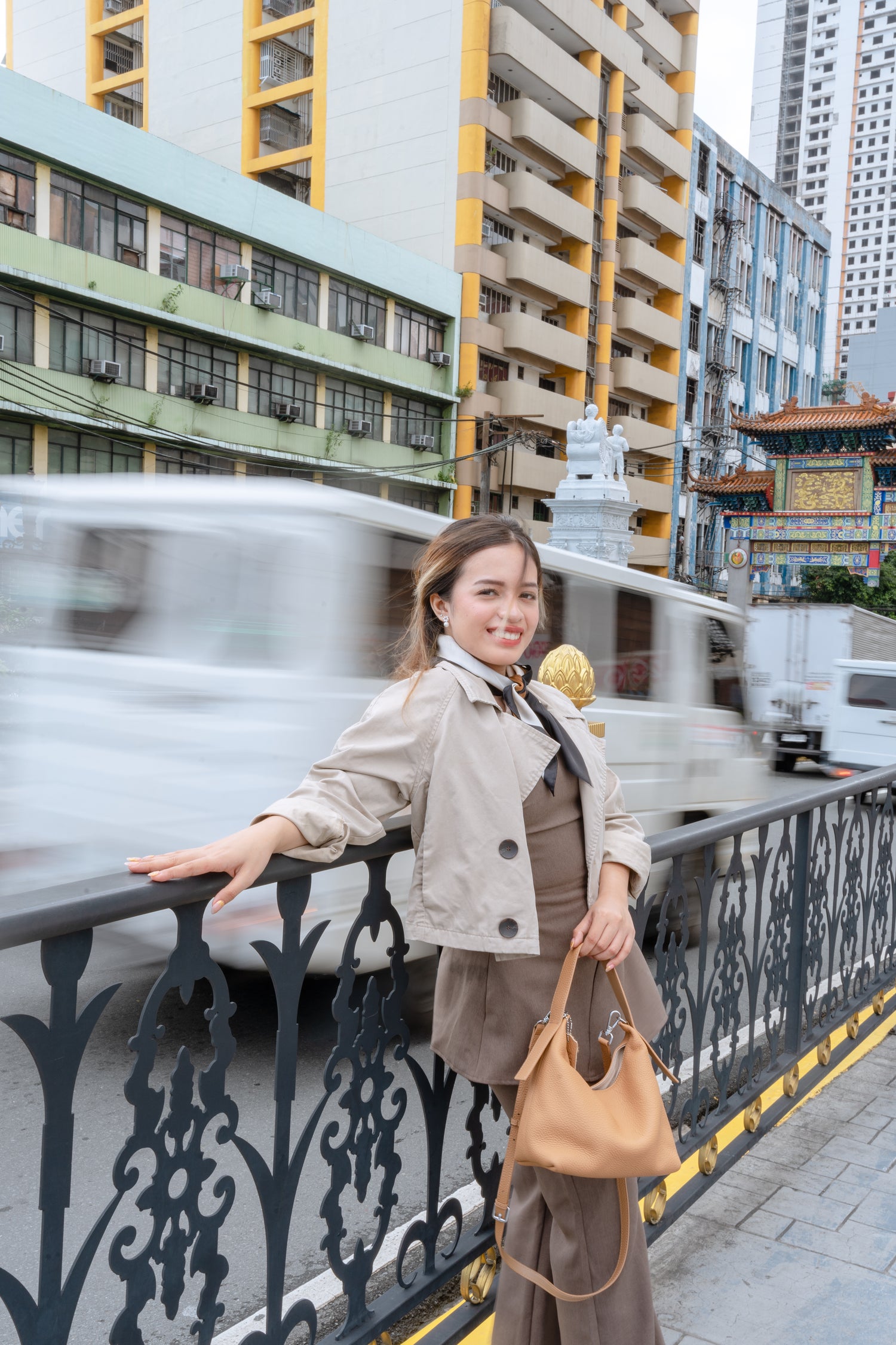 Woman standing on a city street with buildings, carrying a minimalist, quiet luxury bag, and a blurred vehicle in the background